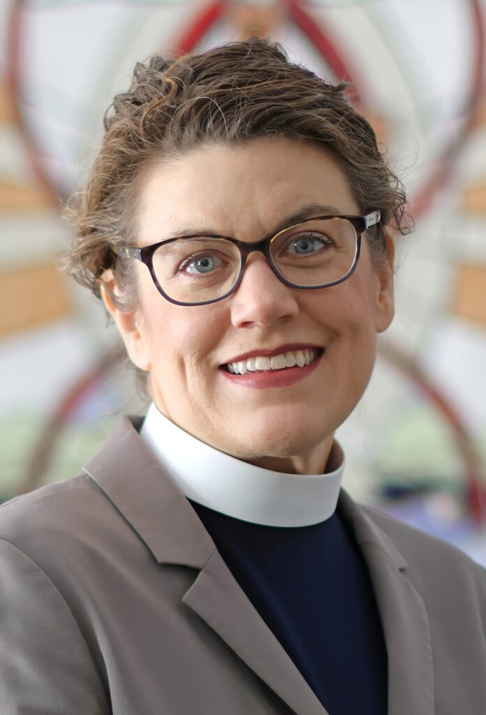 Woman with brown hair pulled back, glasses, clerical collar, and brown dress jacket